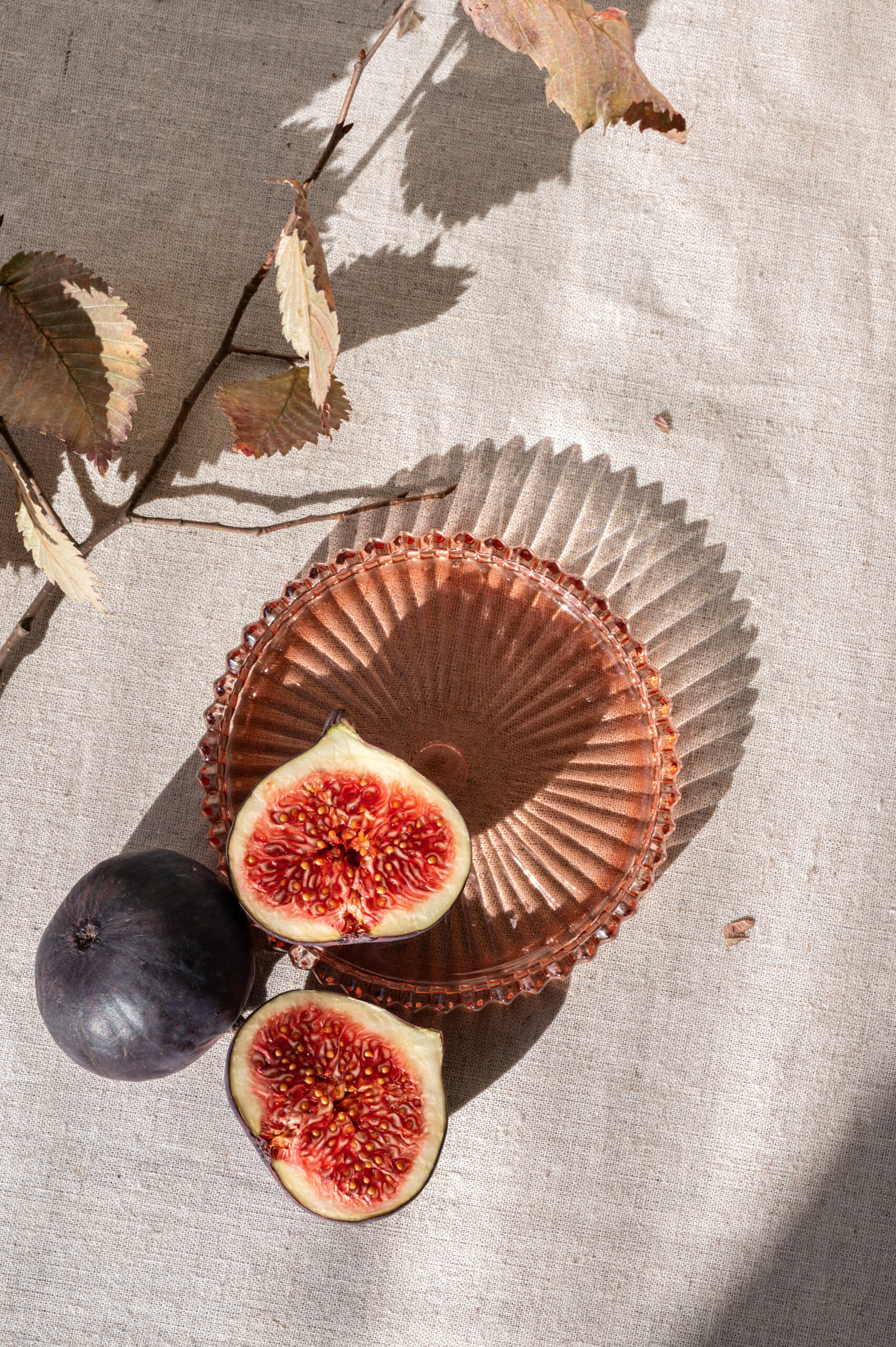 Aesthetic minimal autumn food still life. Whole and cut fig fruits on neutral beige linen tablecloth background with plate, dried leaves and harsh sunlight shadows.
